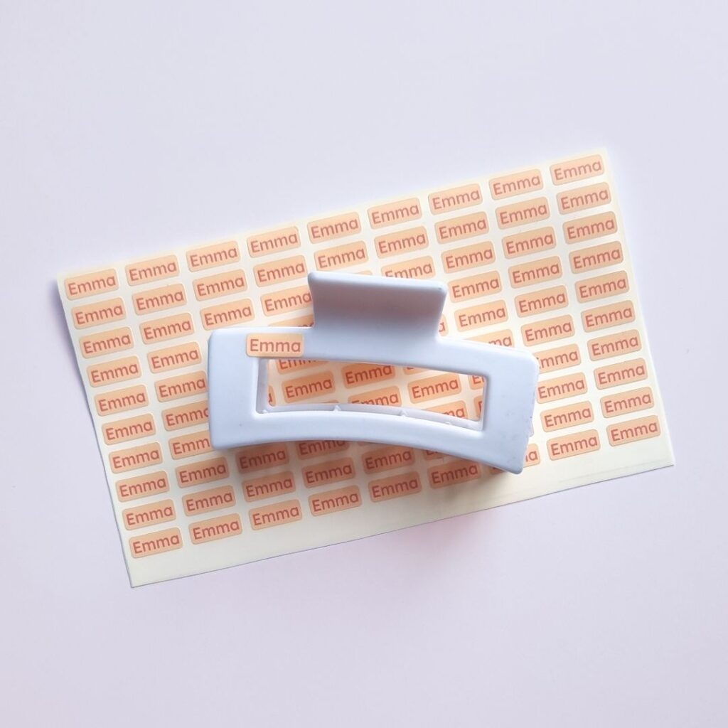Mini name labels in orange with the name emma, placed under and attached to a white hair clip