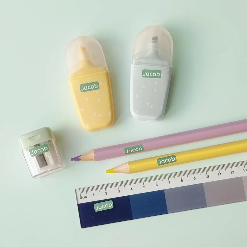 School supplies labeled with green name stickers, including pencils, erasers, a sharpener, and a ruler