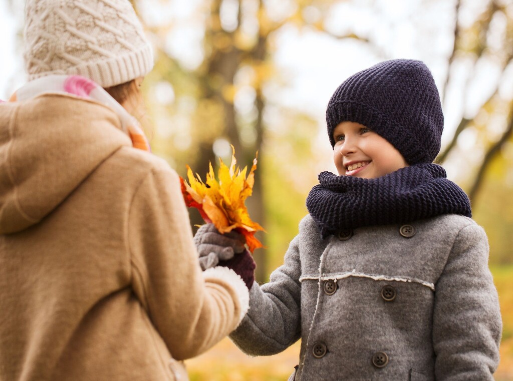 Two children bundled up in winter coats and hats, exchanging vibrant autumn leaves in a park