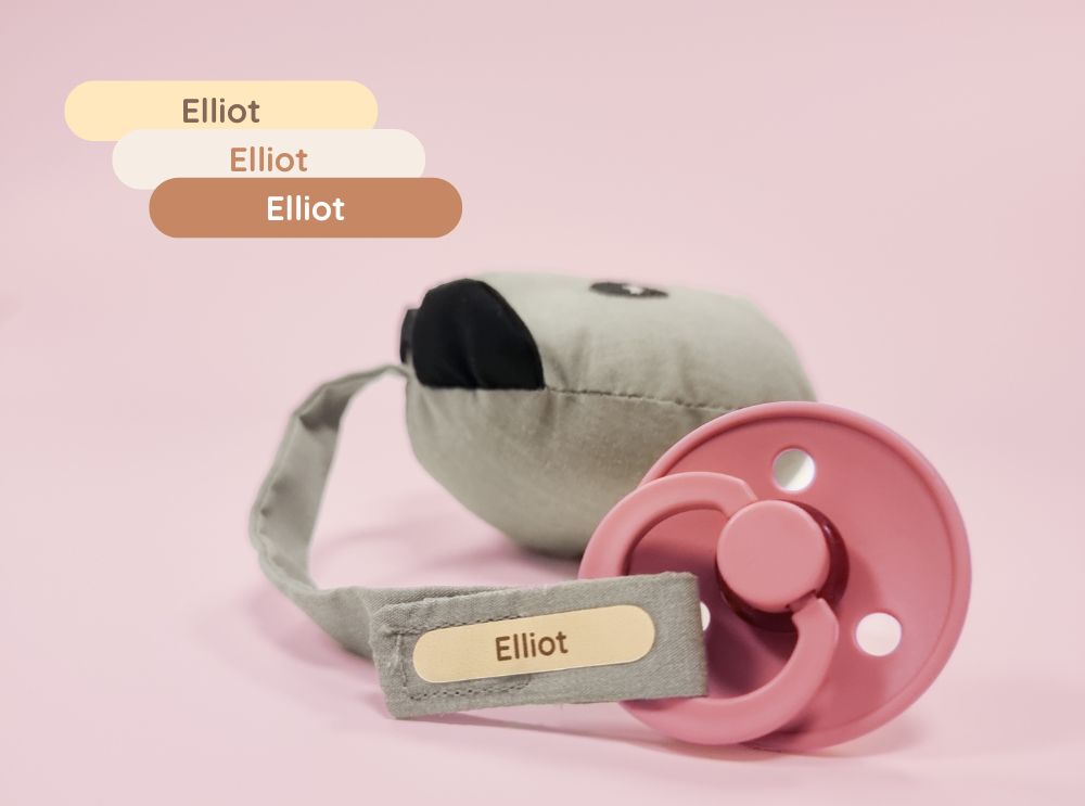 Pink dummy with a personalised name label reading 'elliot' on a fabric strap.