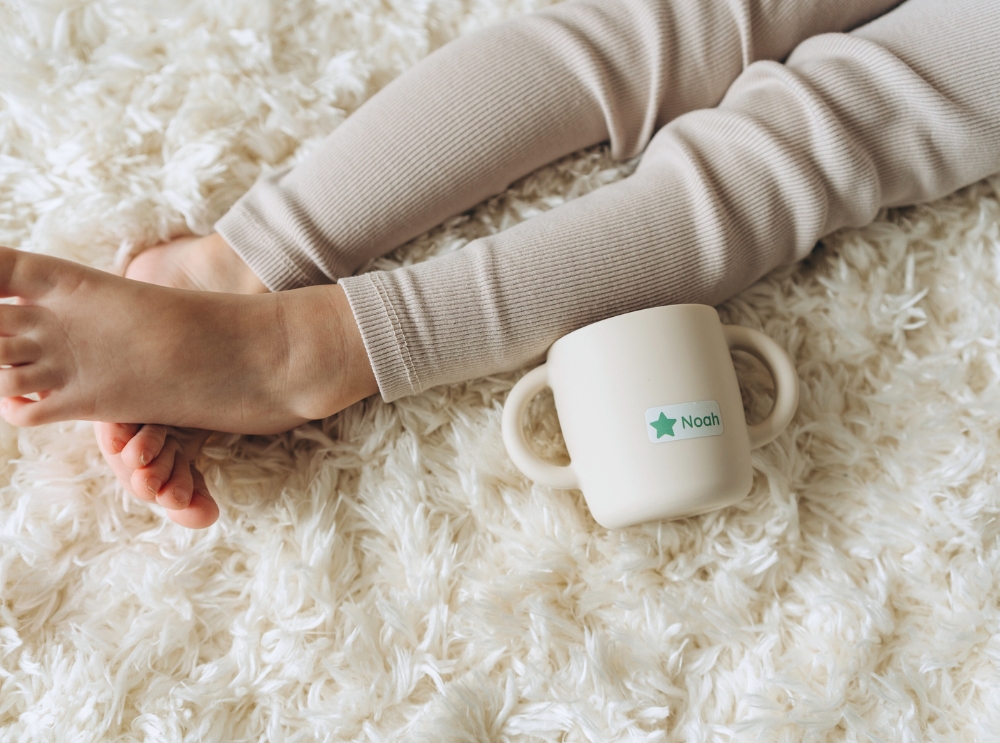 Toddler cup with a personalised name label on a soft, fluffy rug next to a child's legs.