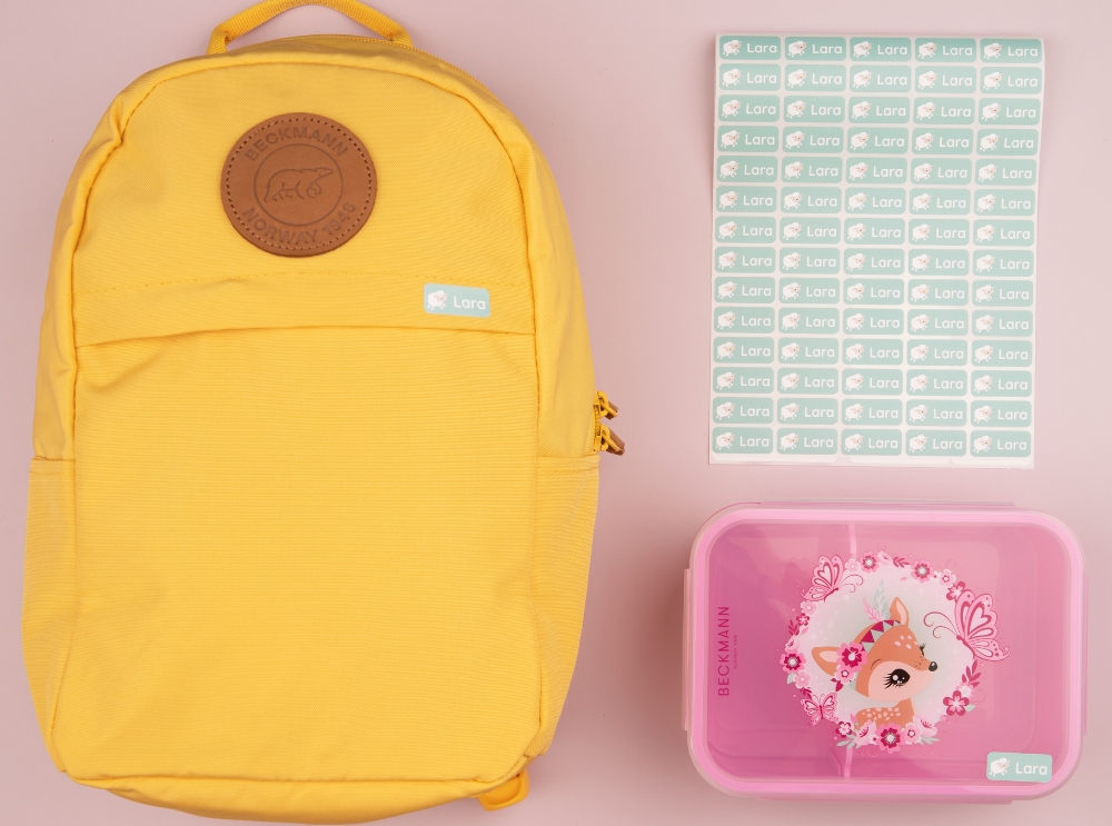 Yellow backpack, pink lunchbox, and a sheet of green name labels arranged on a pink background