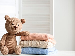 Knitted teddy bear sitting next to a stack of folded baby clothes in soft pastel colors