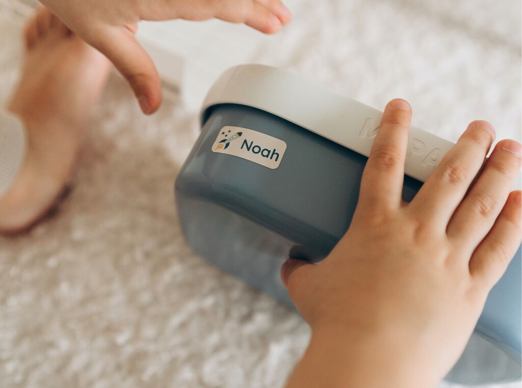 Close-up of a child’s hands holding a blue lunchbox labelled with a personalised name sticker featuring a rocket and the name noah