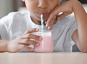Child sipping a pink smoothie from a glass with a striped straw at a table.