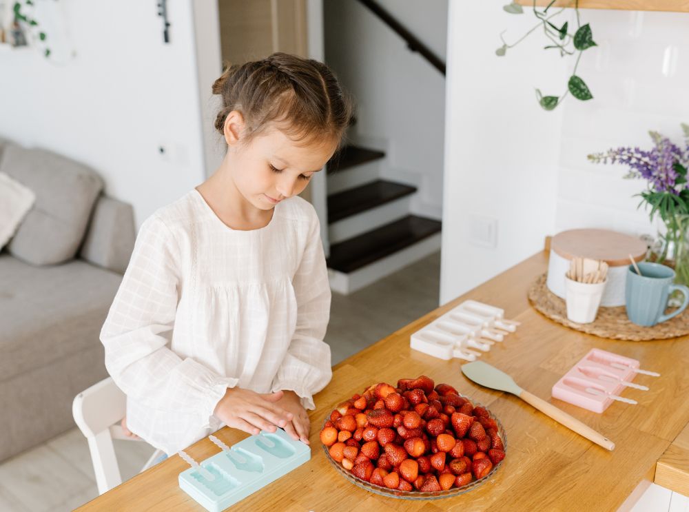 Young girl preparing strawberries at a kitchen table with lolly moulds nearby, getting ready to make homemade frozen treats.