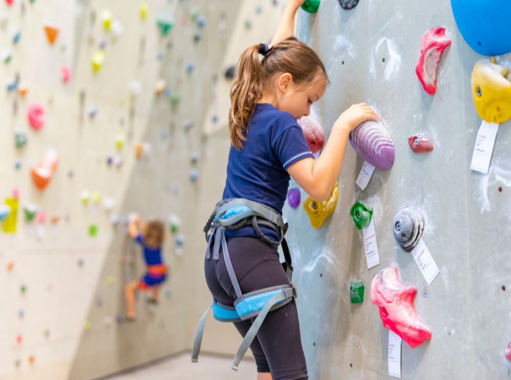 Child climbing an indoor rock wall, wearing a harness and reaching for colourful holds.