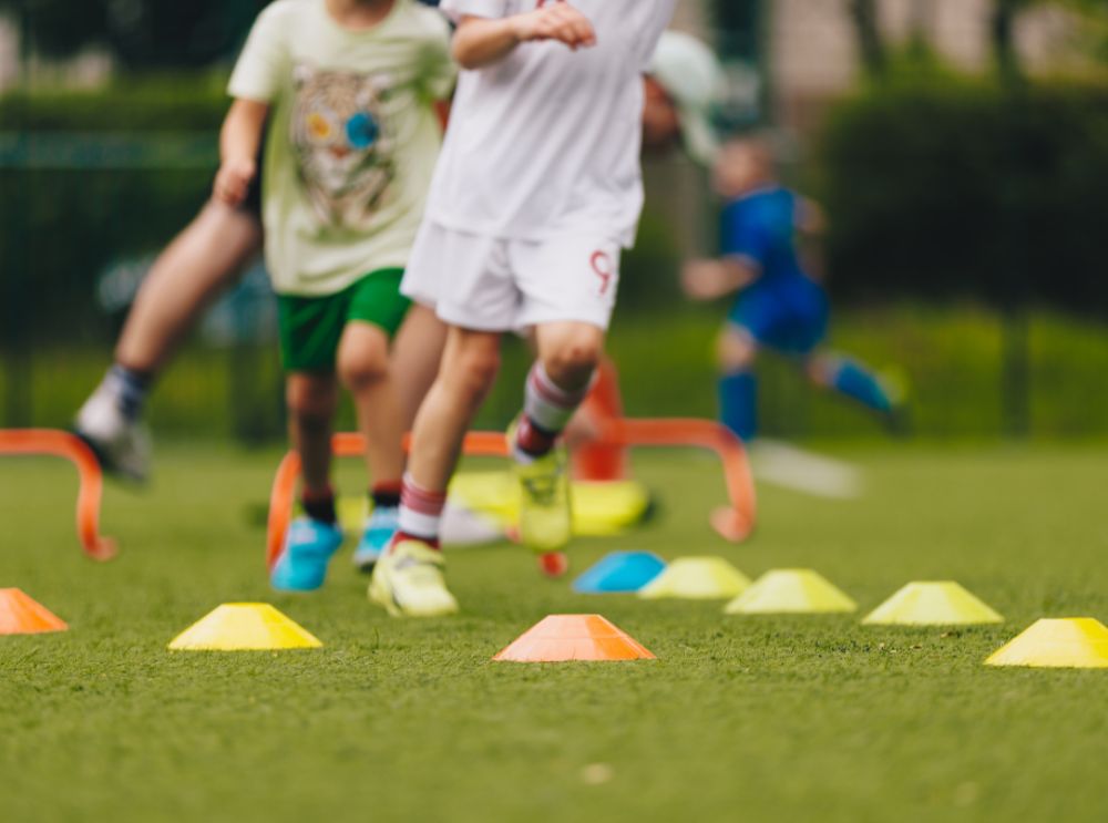 Children running through football training cones and hurdles on a pitch.