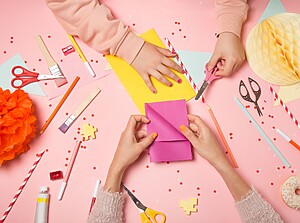 Hands crafting colorful birthday invitations with paper, scissors, and decorations on a pink table.