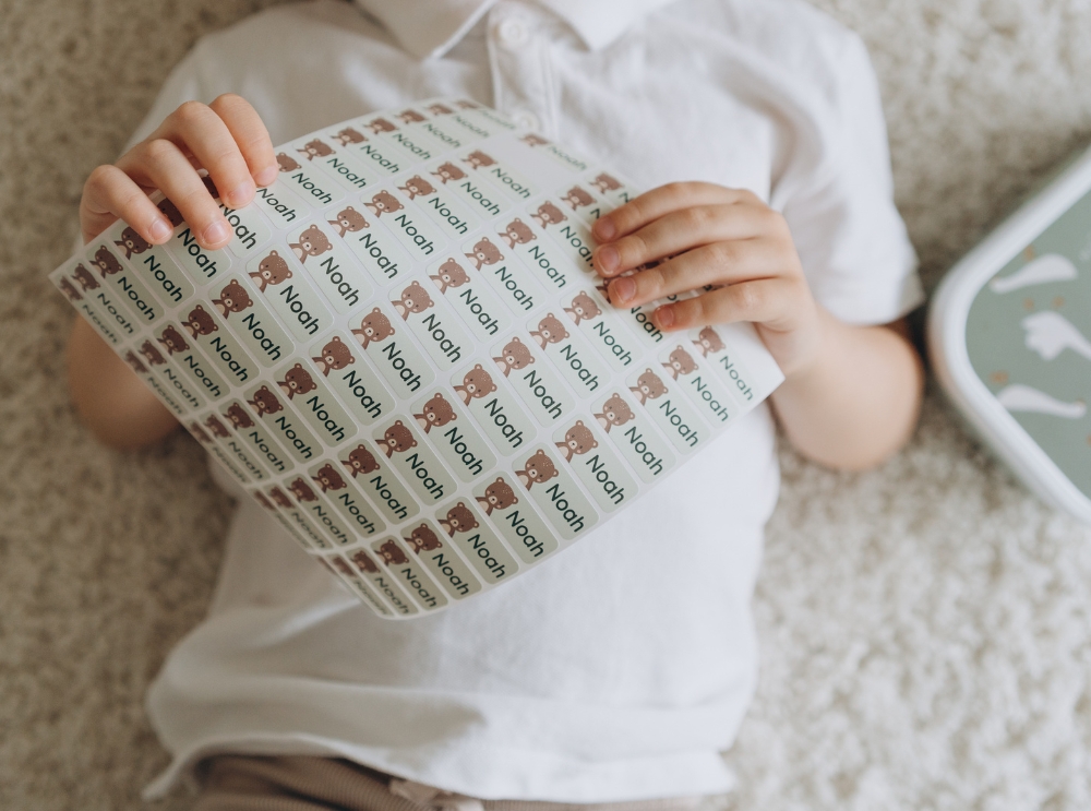 Child holding a full sheet of personalised name labels featuring a bear icon and the name noah