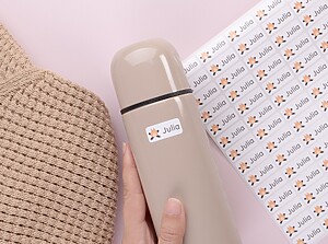Hand holding a beige water bottle labeled with a white name sticker featuring a bonfire and the name Julia, next to a matching sticker sheet