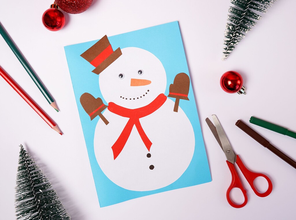 Snowman christmas craft made from cut-out paper shapes, surrounded by pencils and scissors.