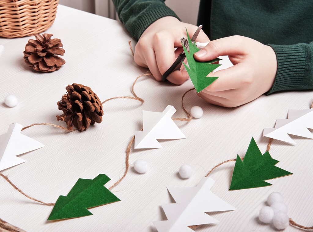 Child crafting a christmas garland with paper trees, pinecones and twine for a cosy holiday decoration.