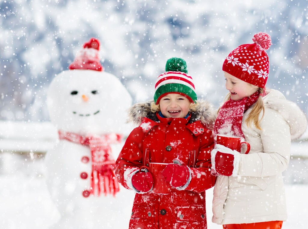 Snow much fun: the best winter activities for kids 2 Two kids standing in front of a snowman, warmly dressed and holding cups of hot cocoa while snow is falling.