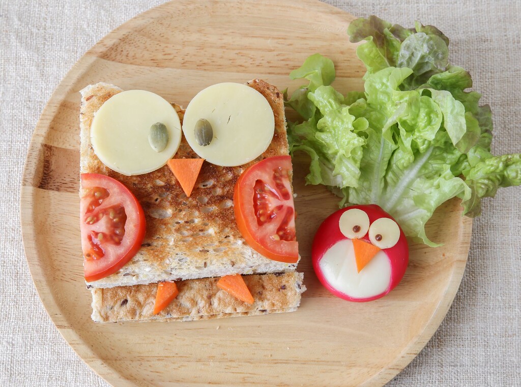 Owl-shaped children’s snack made from toast, cheese, tomato, carrot, with a little egg chick on a wooden plate.