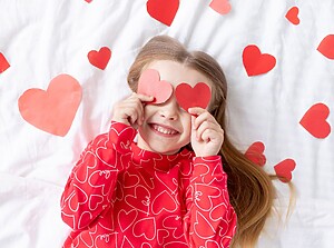 Smiling child holding red paper hearts over their eyes, lying on a white background with heart decorations.