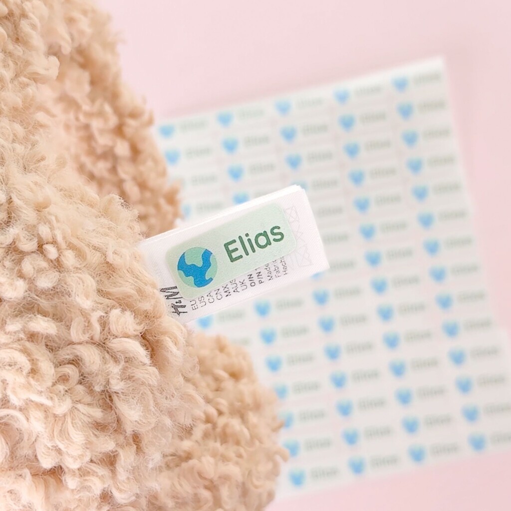 Close-up of a personalised name label featuring the name elias and a globe icon on a clothing label, with a matching sticker sheet behind.