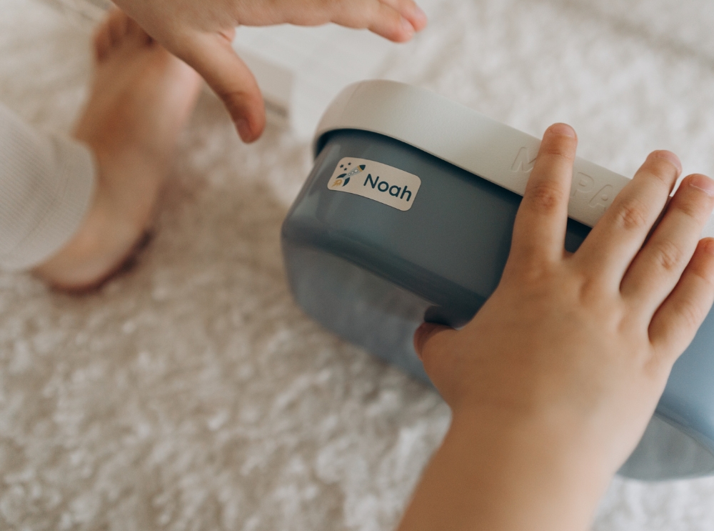Close-up of a child’s hands holding a blue lunchbox labeled with a personalized name sticker featuring a rocket and the name noah