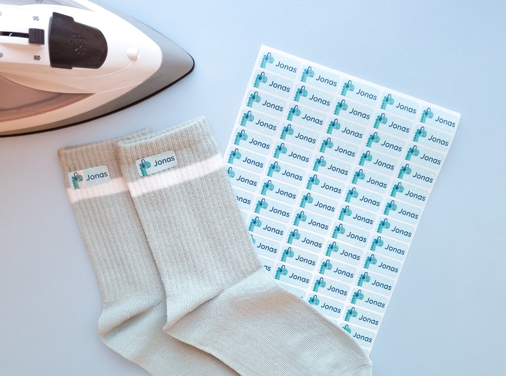 How to apply iron-on labels: a simple guide for long-lasting results 2 Sheet of personalised iron-on namelabels with a dinosaur wearing headphones, next to labelled socks and an iron